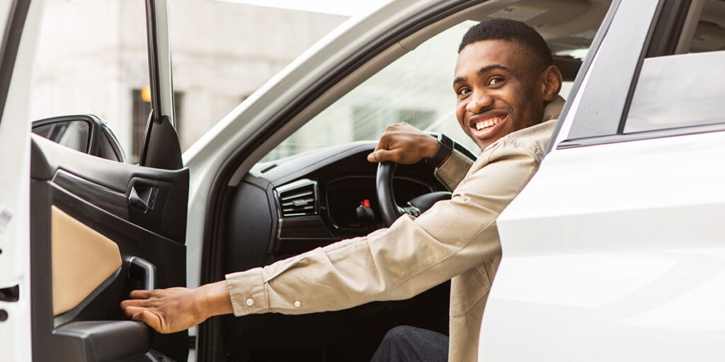 Smiling man sitting in the driver’s seat of a car, holding the door open.