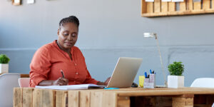 balancing short term and long term financial goals Woman reviewing notes and working on a laptop at a desk in a bright home office